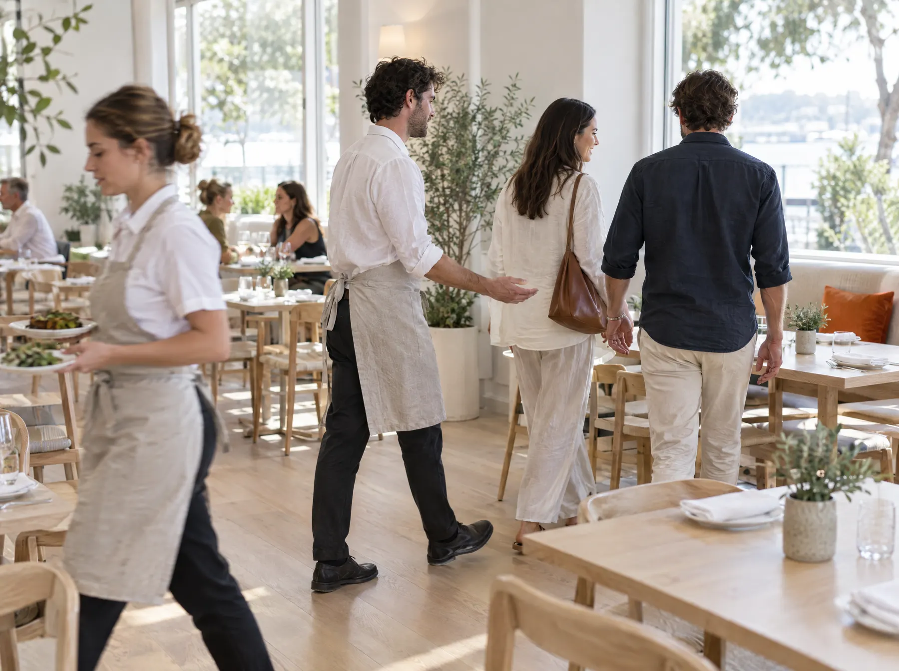 A host walks two guests to their table while a server carries plates through a bright dining room.